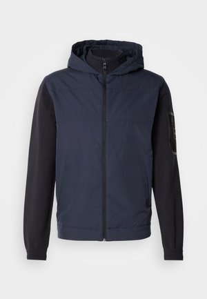 Chaqueta azul marino con capucha; combina una tela lisa con una manga texturizada oscura. Cuenta con una cremallera frontal y bolsillos laterales.