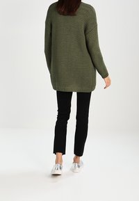 Suéter de punto verde oliva con un corte holgado y mangas largas, combinado con jeans negros ajustados y zapatos plateados de pala.