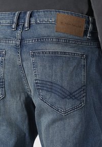 Modré džínové kalhoty z denimu s koženou nášivkou značky na pasu. Mají zadní kapsy s obloukovým šitím a vybledlou texturou.