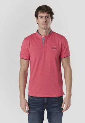 Polo rosa con pequeños puntos azules y blancos, manga corta, tapeta con dos botones y detalles en el cuello azul. Se lleva con vaqueros azul oscuro.