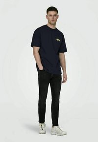 T-shirt de algodão azul-marinho com mangas curtas, apresentando um logótipo amarelo no peito. Usada com jeans pretos e ténis brancos.