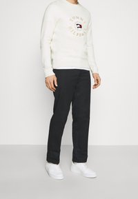 Weißer Sweatshirt mit Logo-Druck in der Mitte, kombiniert mit schwarzen Hosen und weißen Turnschuhen. Das Outfit hat ein lässiges Design und einfarbige Farben.