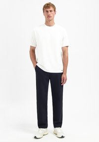 Witte korte mouwen t-shirt in combinatie met navy broek en lichtgekleurde sneakers. De stof lijkt soepel; de pasvorm is ontspannen.