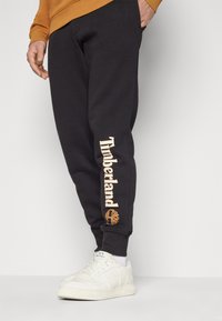 Svarta joggingbyxor med "Timberland"-text i ljusbrunt på vänstra benet, i kombination med vita sneakers, vilket visar en avslappnad stil.