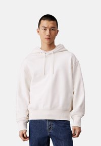 Felpa bianca realizzata in tessuto morbido con vestibilità comoda, cappuccio con cordini, polsini e orlo a costine, abbinata a jeans blu.