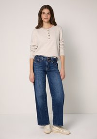 Beżowa koszulka henley z długim rękawem z trzema guzikami, zestawiona z szerokimi niebieskimi dżinsami i beżowymi tenisówkami. Prosty design, lekka faktura, casualowy krój.