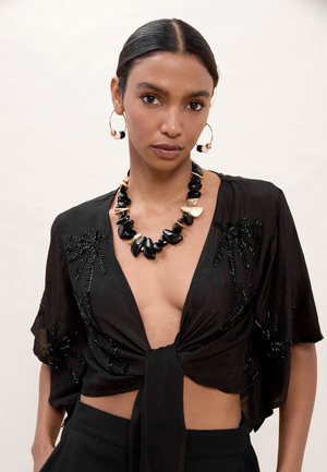 Mujer con top corto negro atado con detalles bordados, un gran collar grueso negro y dorado, y pendientes de aro dorados con cuentas.