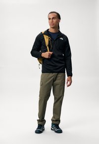 Chaqueta negra de pullover con media cremallera, pantalones cargo oliva, zapatillas deportivas azules y mochila amarilla. Diseño minimalista, tejido funcional.