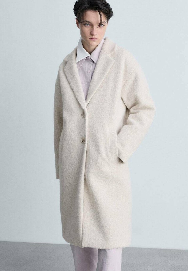 Cappotto di lana color crema con collo a revers, maniche lunghe, due bottoni e tasche laterali. Indossato sopra una camicia chiara.