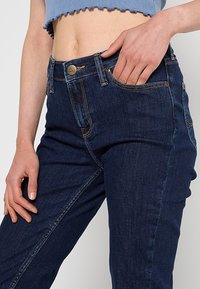Femme portant un jean Lee bleu foncé et un haut court bleu, une main dans la poche et l'autre reposant le long du corps.