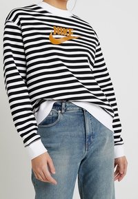 Svart- och vitrandig sweatshirt med rund halsringning, prydd med en orange Nike-logga. Tillsammans med ljusblå denimjeans.