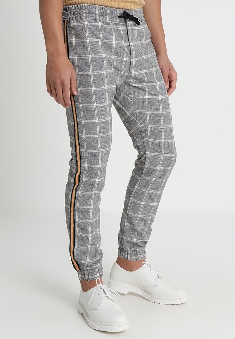 Grijze geruite joggerbroek met een elastische tailleband, zwarte trekkoord en zijstrepen in oranje en zwart. G gedragen met witte sneakers.