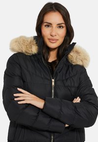 Zwarte pufferjack met een capuchon met bontafwerking, ritsluiting, gewatteerde textuur en lange mouwen. Het model heeft haar armen gekruist en toont de jas.