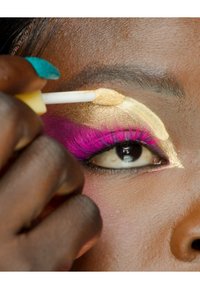 Application d'ombre à paupières dorée sur la paupière, avec de faux cils roses vifs et une main brune tenant un applicateur en éponge.