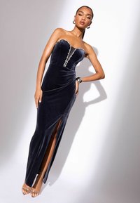 Bešvarkė tamsiai mėlynos aksomo suknelė be petnešų, turinti prigludusį liemenį, puošiasi detale per vidurį ir turi aukštą skeltą iki šlaunies. Matomos skaidrios kulninės šlepetės.