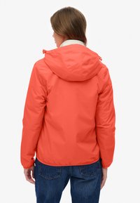 Person iført en koralfarvet hættetrøje med løbesnore, hvid krave synlig og blå jeans, stående med ryggen mod en ensfarvet baggrund.