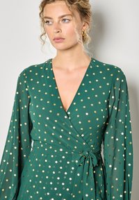 Abito verde avvolgente con pois dorati e bianchi, maniche lunghe, scollatura a V e una cintura legata in vita. Tessuto morbido, caratterizzato da un design fluido.