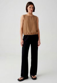 Woman standing wearing a sleeveless brown top, black wide-leg pants, and black platform sandals against a plain light background.