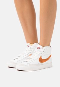 Nike högskosneakers har en vit läderöverdel, orange mocka-swoosh och en grå gummisula med texturerad detaljering.