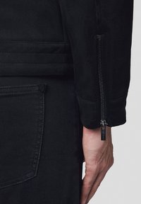 Czarna kurtka o teksturowanym wykończeniu z detalem w postaci zamka błyskawicznego na rękawie. Zbliżenie na rękaw i dłoń, ukazujące materiał i szycie. Gładkie wykończenie.