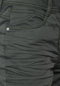 Pantalones de un verde oscuro con una textura arrugada, que cuentan con detalles de costura sutiles y un acento de botón metálico cerca del bolsillo.