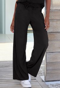 Black wide-leg pants made of smooth fabric with an elastic waistband and drawstring detail, paired with white sneakers.