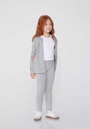 Haut à zip gris, t-shirt blanc et leggings gris. Une fille se tient en baskets blanches. Texture de tissu lisse et design simple.