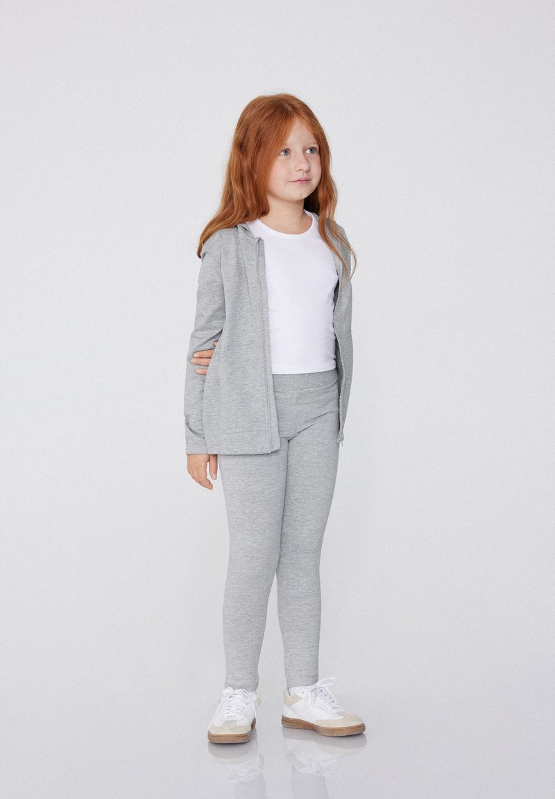Šedá mikina na zip, bílý tričko a šedé legíny. Dívka stojí v bílých teniskách. Hladká textura látky a jednoduchý design.