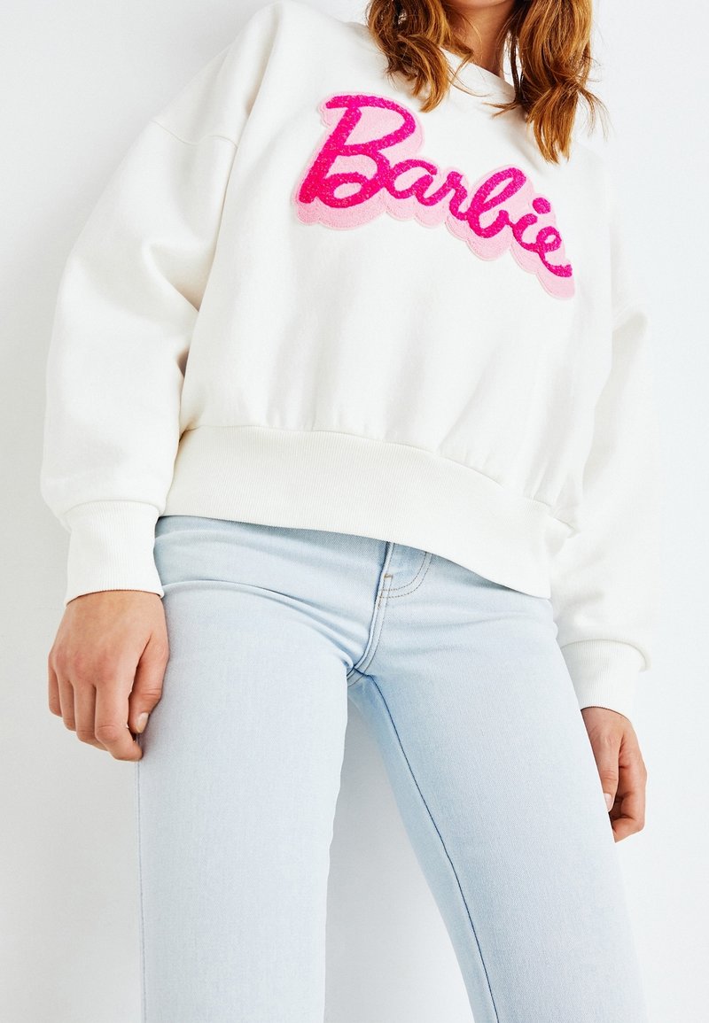 Femme portant un sweat-shirt blanc avec "Barbie" en écriture rose et un jean bleu clair, montrée des épaules jusqu’aux hanches sur fond blanc.