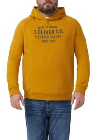 Mężczyzna w musztardowym hoodie z napisem "S. Oliver Co. Elevated Casual Since 1969", w połączeniu z niebieskimi dżinsami, na białym tle.