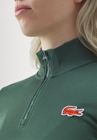 Zelený žebrovaný zip-up top s vysokým límcem a oranžovou vyšívanou logem na prsou, s kovovým zipem.