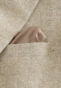 Beiger strukturierter Stoff mit einem Taschentuch aus weichem braunen Seidenmaterial, das teilweise in der linken Tasche eines hellen Blazers sichtbar ist.