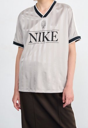 Osoba nosząca jasnoszarą sportową koszulkę Nike z czarnym obszyciem oraz dużym czarnym logo Nike i tekstem z przodu, w połączeniu z ciemnymi spodniami.