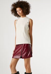 Göndör hajú nő, akinek ujjatlan, lyukacsos részletekkel ellátott fehér topja és fényes piros bőr mini szoknyája van, egy fehér háttér előtt áll.