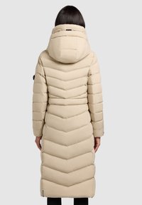 Bézs hosszú steppelt kabát cikkcakk mintázattal, magas nyakú kapucnival és elasztikus mandzsettákkal. Oldalsó logórészlettel és középmankós hosszúsággal.
