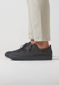 Chaussures en suede noir avec un bout arrondi, un laçage à l'avant et une semelle en caoutchouc. Portées avec un pantalon légèrement fuselé de couleur claire.