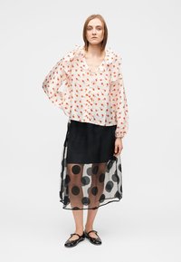 Blouse légère et transparente à fleurs avec un col à volants en pêche et rouge, superposée sur une jupe noire opaque à pois, à la silhouette fluide.