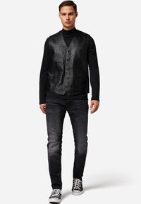 Schwarze Lederweste mit V-Ausschnitt, sechs Knöpfen und zwei Vorderaschen, getragen über einem schwarzen Langarmshirt und dunklen Jeans mit Sneakers.