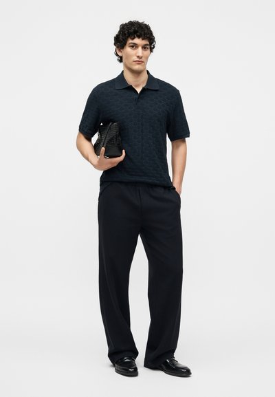 Άνδρας που φοράει μαύρο πόλο με υφή και φαρδιά μαύρα παντελόνια, κρατώντας μαύρη τσάντα, στέκεται μπροστά σε απλό ανοιχτό φόντο.