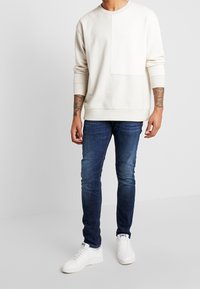 Hombre con sudadera de manga larga color crema, jeans pitillo azul oscuro, zapatillas blancas y tatuajes visibles en el brazo, contra un fondo liso y claro.