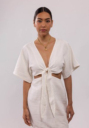 Haut en lin blanc à manches courtes et devant noué, associé à une jupe ajustée. Un collier en or avec un pendentif accentue l'ensemble.