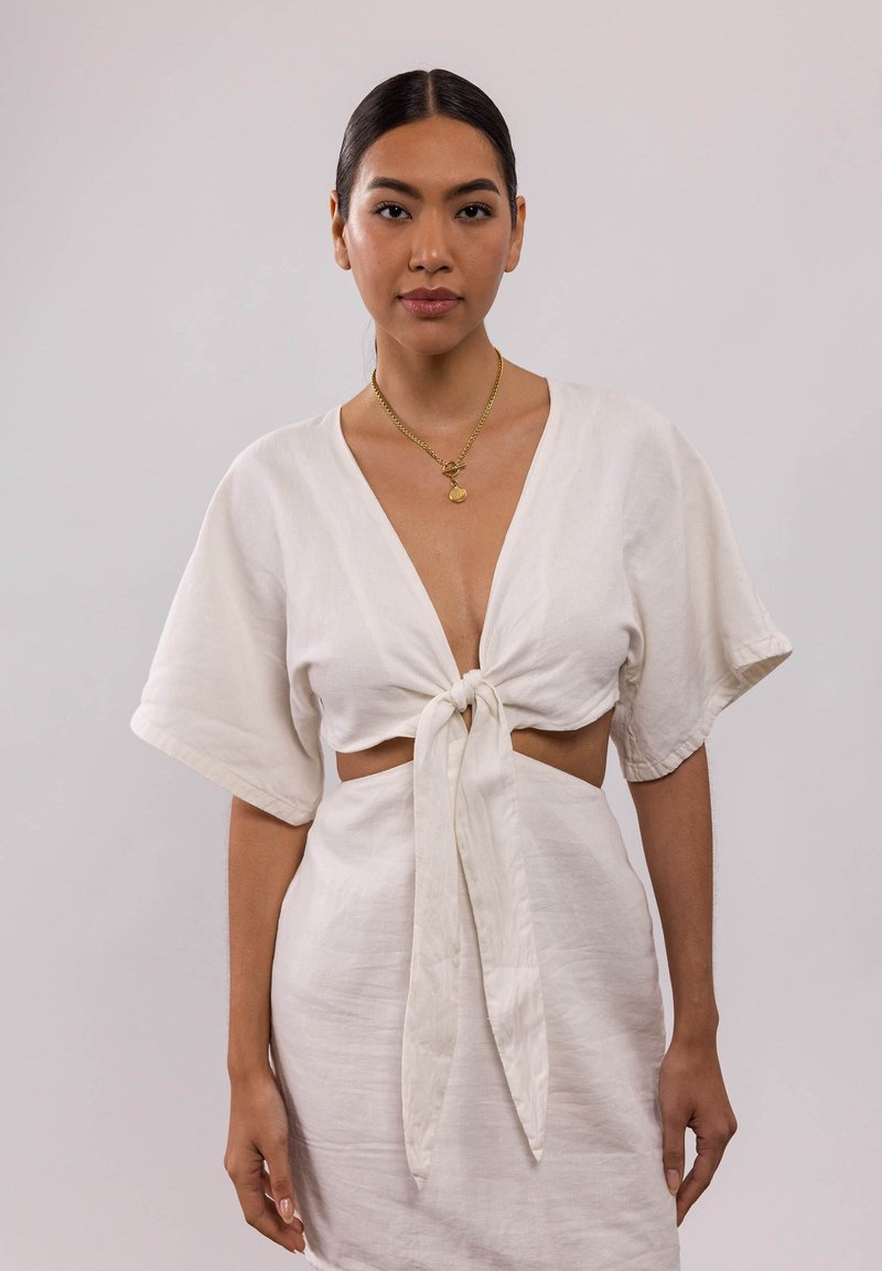 Weißes Leinenoberteil mit kurzen Ärmeln und einer geknoteten Vorderseite, kombiniert mit einem taillierten Rock. Eine goldene Halskette mit einem Anhänger akzentuiert das Outfit.