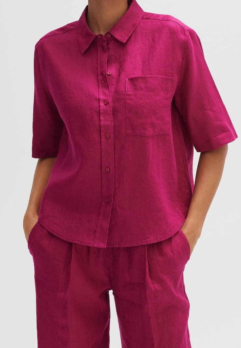 Femme portant une chemise boutonnée en lin magenta à manches courtes et un pantalon taille haute assorti, les mains dans les poches.