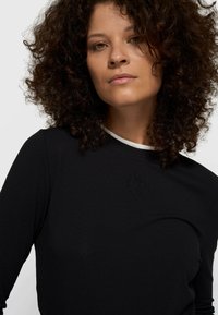 Top negro de manga larga con escote redondo y ribete blanco a contraste, con detalle de logo en relieve en el pecho. Textura de tela suave.