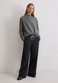 Grauer Pullover mit hohem Kragen, kombiniert mit lockeren schwarzen Jogginghosen mit Kordelzug und weißen Seitenstreifen. Modell steht vor einem neutralen Hintergrund.