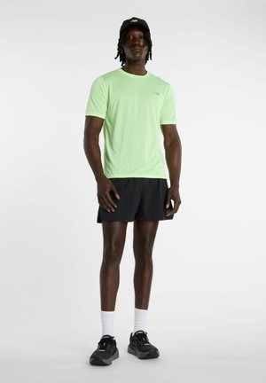 Uomo in piedi che indossa una maglietta verde chiaro a maniche corte, pantaloncini neri, cappello nero, calzini bianchi e scarpe sportive nere su uno sfondo semplice.