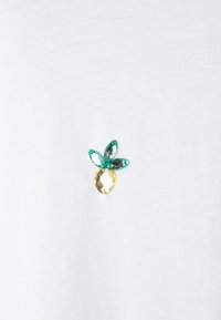 T-shirt blanc comportant une broche décorative multicolore en forme de fruit, avec des accents de joyaux verts et jaunes. Texture de tissu lisse.