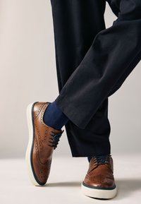 Braune Brogues aus Leder mit gestanzten Details, blauen Schnürsenkeln und weißen Gummisohlen, kombiniert mit dunklen Hosen und marineblauen Socken.