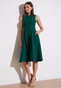 Rochie verde, fără mâneci, până la genunchi, cu un corset ajustat, fustă evazată și buzunare laterale. Material neted, de o culoare solidă.