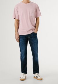 Ljust rosa bomullströja med bröstficka, kombinerad med mörkblå denimjeans och vita sneakers med svarta detaljer.